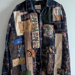 May include: A patchwork flannel shirt in navy blue, brown, and cream. The shirt features various fabric patches with floral and geometric patterns. The shirt is hanging on a blue hanger against a white wall.