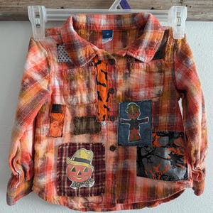 May include: A red and orange plaid flannel shirt with various Halloween-themed patches sewn on. The patches include a pumpkin, a scarecrow, a witch, and a ghost. The shirt has a button-up closure and long sleeves.