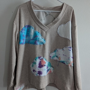 May include: A beige v-neck sweatshirt with cloud appliqués. The clouds are in shades of blue, white, purple, and orange. The sweatshirt has a tag that says "Wear You Love". The brand name "Orvis" is visible on the inside of the collar.
