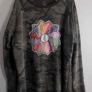 May include: A camouflage-patterned sweatshirt with a colorful, flower-shaped appliqué. The appliqué is made of various fabric scraps in shades of pink, blue, yellow, and patterned fabrics. The sweatshirt has a crew neck and long sleeves.
