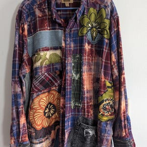 May include: A patchwork button-down shirt with a plaid pattern in shades of blue, red, and orange. The shirt features various fabric patches with floral and striped designs, and a small denim pocket. The shirt is hanging on a blue hanger.