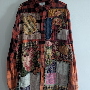 May include: A patchwork button-down shirt with a red and black plaid pattern, featuring floral and denim patches. The shirt is hanging on a white hanger against a white wall and a wooden plank.