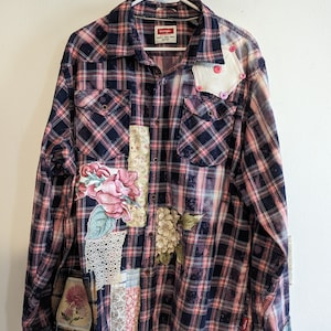 May include: A black and pink plaid flannel shirt with various fabric patches and floral embroidery. The shirt has a button-up front and long sleeves. The label on the shirt reads "Wrangler".