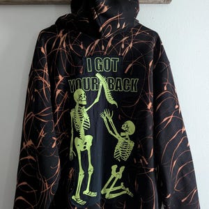 May include: Black hoodie with a neon green skeleton graphic and the text "I GOT YOUR BACK." The hoodie has an orange splatter design. The graphic shows two skeletons, one standing and one kneeling.
