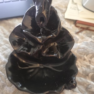 May include: A black ceramic incense burner featuring a tiered, leaf-shaped design. The structure consists of multiple overlapping, curved petal forms that create a cascading effect, with a small opening at the top for incense placement.
