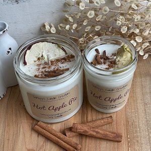 May include: Two soy candles in glass jars with lids. The candles are labeled "Hot Apple Pie" and feature a cinnamon stick and apple slice on top. The candles are on a wooden surface with cinnamon sticks scattered around them.