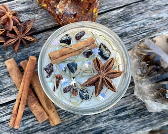 Könnte beinhalten: Eine weiße Kerze mit Zimtstangen, Sternanis und schwarzen Kristallen, die in das Wachs eingebettet sind. Die Kerze befindet sich in einem durchsichtigen Glasgefäß und ist von anderen natürlichen Elementen umgeben, darunter Zimtstangen, Sternanis und ein Stück Bernstein.