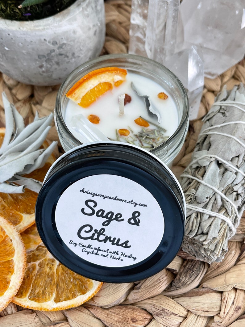 May include: A white soy candle in a glass jar with a black lid. The candle is infused with sage, citrus, crystals, and herbs. The jar has a white label with the text "Sage & Citrus" and "Soy Candle infused with Healing Crystals and Herbs".