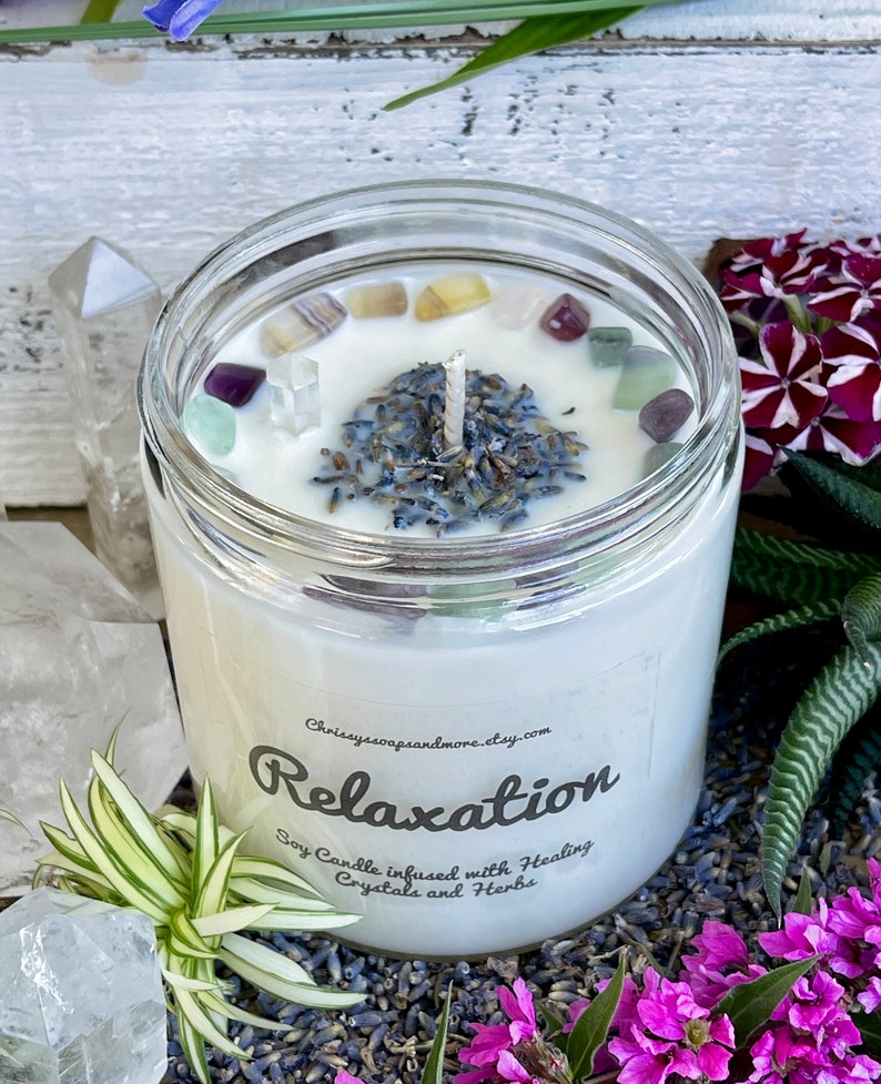 May include: A white soy candle in a clear glass jar with a label that reads "Relaxation Soy Candle infused with Healing Crystals and Herbs". The candle is surrounded by lavender flowers, crystals, and greenery.