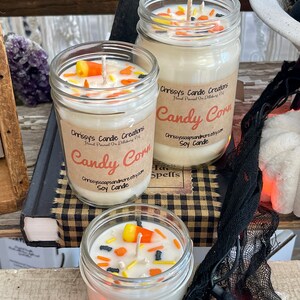 May include: Three glass jars filled with white soy candles decorated with candy corn and black sprinkles. The jars have brown labels with the text "Chrissy's Candle Creations" and "Candy Corn" in a handwritten font. The candles are on a checkered surface.