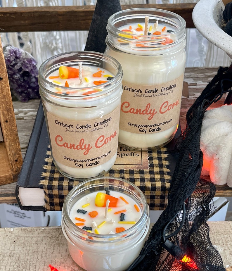 May include: Three glass jars filled with white soy candles decorated with candy corn and black sprinkles. The jars have brown labels with the text "Chrissy's Candle Creations" and "Candy Corn" in a handwritten font. The candles are on a checkered surface.