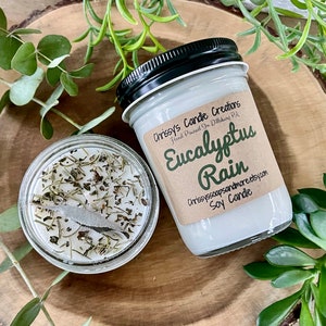 May include: Two soy candles in glass jars. One jar is open, showing dried herbs on the white wax. The other jar has a black lid and a label that reads "Eucalyptus Rain" and "Soy Candle". The candles are on a wooden surface.
