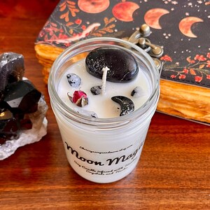 May include: A white candle in a glass jar with a black stone, a dried rose, and black crescent moon charms. The candle is labeled "Moon Magic" and is infused with crystals and herbs.