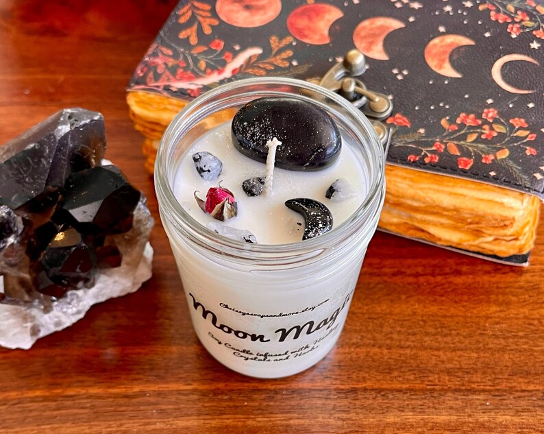 May include: A white candle in a glass jar with a black stone, a dried rose, and black crescent moon charms. The candle is labeled "Moon Magic" and is infused with crystals and herbs.