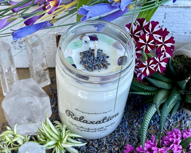 May include: A white soy candle in a clear glass jar with a lavender and crystal top. The candle is labeled "Relaxation Soy Candle Infused with Healing Crystals and Herbs".