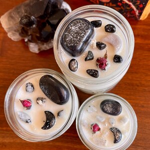 May include: Three glass candle jars with white wax and black stones. The jars are filled with white wax and black stones, and each jar has a small red rose bud.