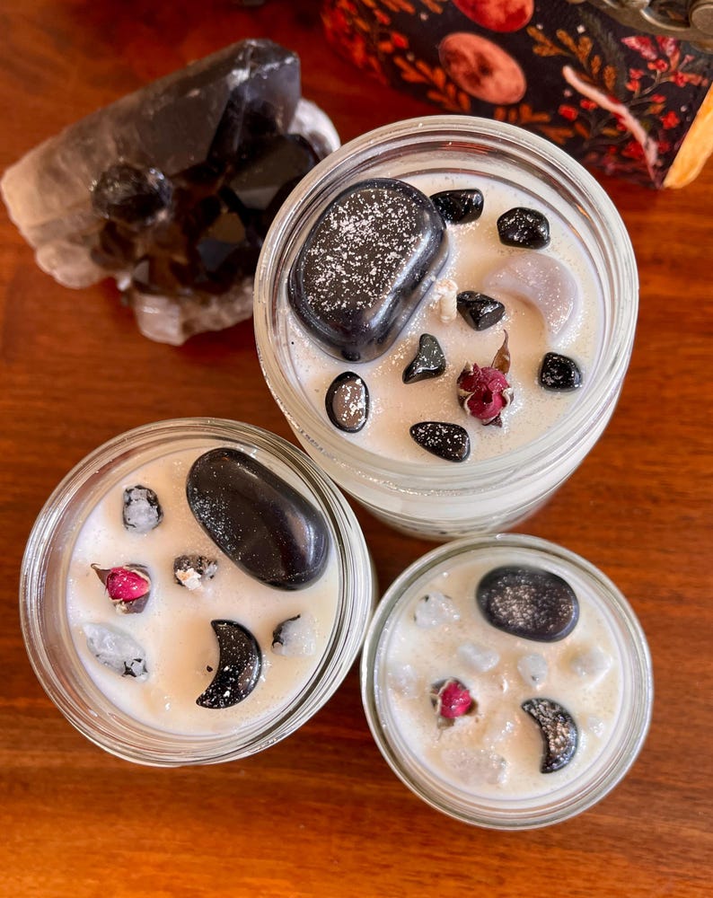 May include: Three glass candle jars with white wax and black stones. The jars are filled with white wax and black stones, and each jar has a small red rose bud.