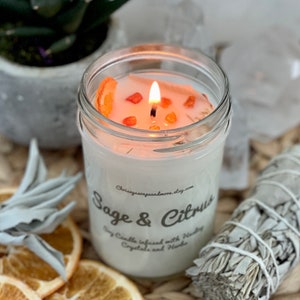 May include: A white soy candle in a glass jar with a white label that reads "Sage & Citrus Soy Candle infused with Healing Crystals and Herbs" and a website address. The candle is lit and has orange slices and orange crystals on top. The candle is surrounded by dried sage leaves and orange slices.