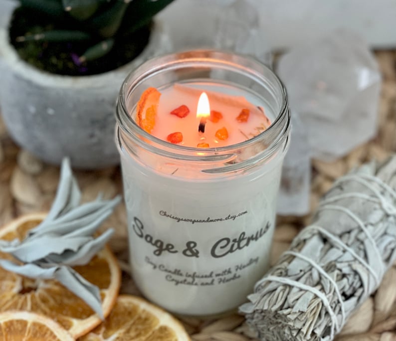 May include: A white soy candle in a glass jar with a white label that reads "Sage & Citrus Soy Candle infused with Healing Crystals and Herbs" and a website address. The candle is lit and has orange slices and orange crystals on top. The candle is surrounded by dried sage leaves and orange slices.