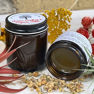 May include: Two brown glass jars with black lids, one open, filled with a salve. The labels read "Arnica, Comfrey & Cayenne Salve" and "Net Wt 4 oz." Dried flowers and herbs surround the jars on a white surface.