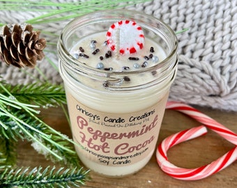 May include: A soy candle in a glass jar, labeled "Peppermint Hot Cocoa." The candle has a white wax base, topped with sprinkles and a peppermint candy. The jar is surrounded by greenery and a candy cane.