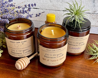 Puede incluir: Tres frascos de vidrio ámbar con tapas que contienen velas de cera de abeja 100% orgánica. Las velas están etiquetadas como "Hint of Natural Honey", "Lavender" y "100% Organic Beeswax Candle". Un cucharón de miel de madera está en primer plano.