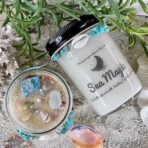 May include: Two glass jars of Sea Magic candles with black lids, adorned with turquoise beads and seashells. The candles are filled with white wax and crystals. The text "Sea Magic" is printed on the front of the candle jars.
