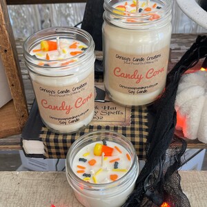 May include: Three glass jars with white soy candles topped with candy corn and sprinkles. The jars have labels that say "Chrissy's Candle Creations" and "Candy Corn".
