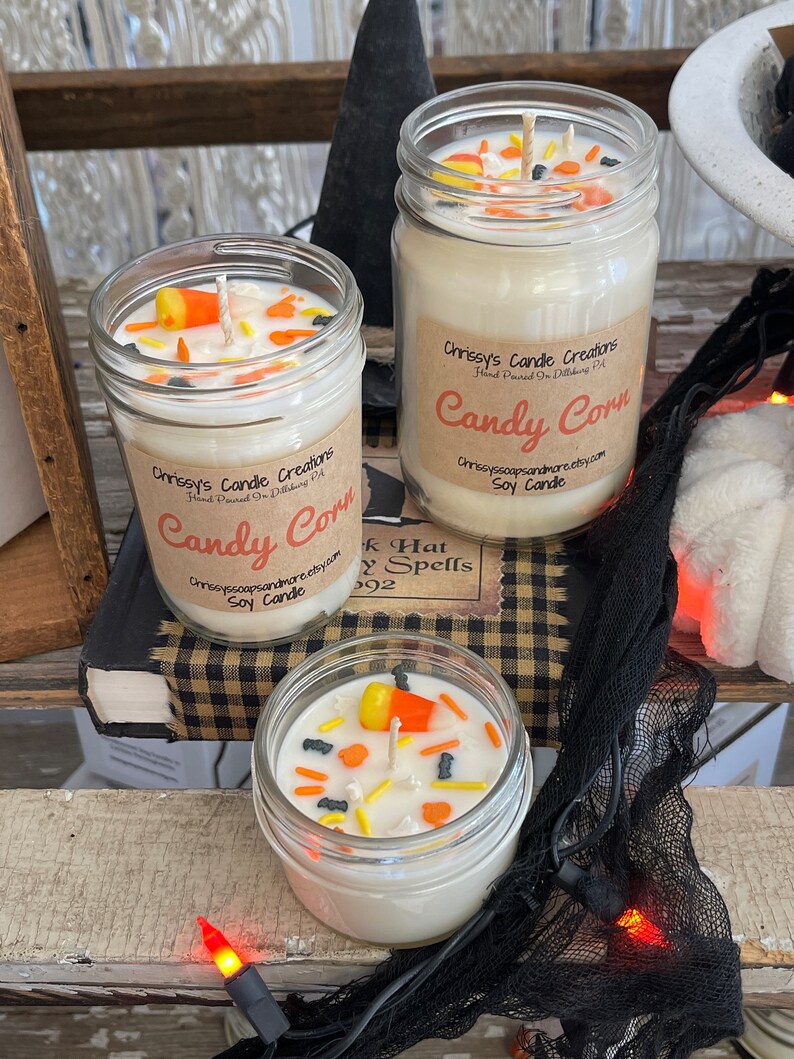 May include: Three glass jars with white soy candles topped with candy corn and sprinkles. The jars have labels that say "Chrissy's Candle Creations" and "Candy Corn".