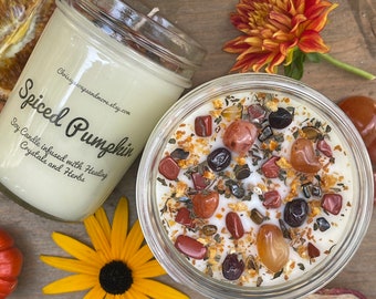 May include: A glass jar candle with a white soy wax base and a label that reads "Spiced Pumpkin Soy Candle infused with Healing Crystals and Herbs". The candle is surrounded by orange and red crystals, dried orange peel, and green herbs.