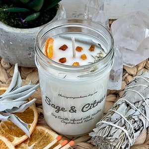 May include: A white soy candle in a glass jar with a sage and citrus scent. The candle is decorated with dried orange slices, amber crystals, and herbs. The jar has a label that reads "Sage & Citrus" and "Soy Candle infused with Healing Crystals and Herbs".