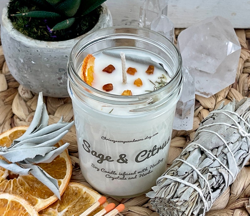 May include: A white soy candle in a glass jar with a sage and citrus scent. The candle is decorated with dried orange slices, amber crystals, and herbs. The jar has a label that reads "Sage & Citrus" and "Soy Candle infused with Healing Crystals and Herbs".