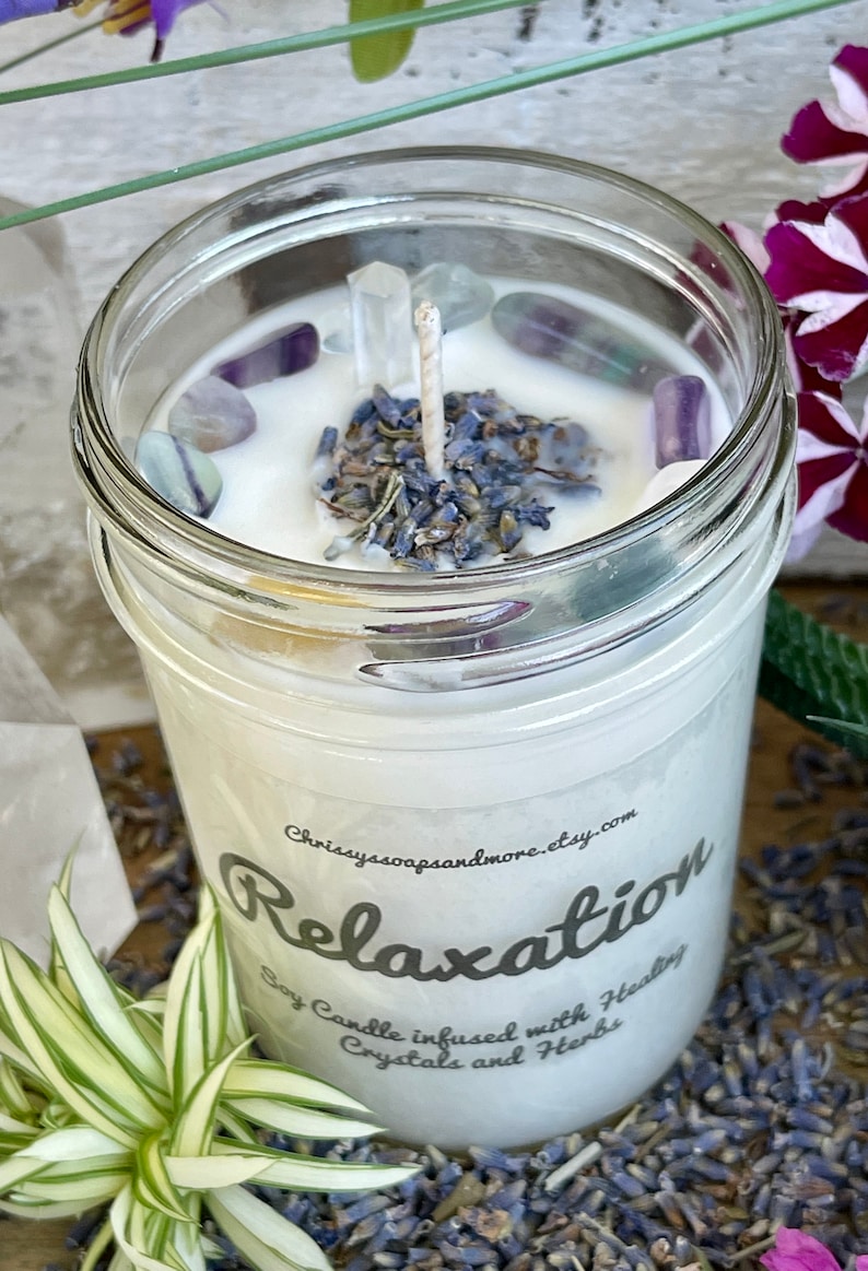 May include: A white soy candle in a clear glass jar with a white label that reads "Relaxation Soy Candle infused with Healing Crystals and Herbs". The candle is topped with dried lavender and clear crystals.