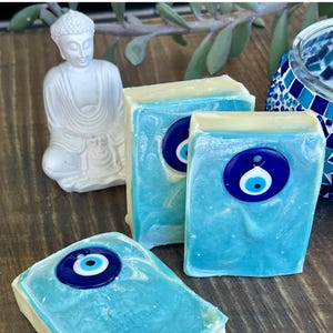 May include: Three rectangular bars of soap with a marbled light blue and white finish. Each bar is embedded with a circular dark blue glass evil eye charm featuring a white and light blue center. The soap bars have a smooth, solid texture.