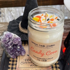 May include: A glass jar candle with a white soy wax base and a candy corn scent. The jar is topped with a wooden wick and decorated with candy corn pieces and sprinkles. The label reads "Chrissy's Candle Creations Hand Poured in Dillsburg PA Candy Corn Chrissyssoapsandmore.etsy.com Soy Candle".