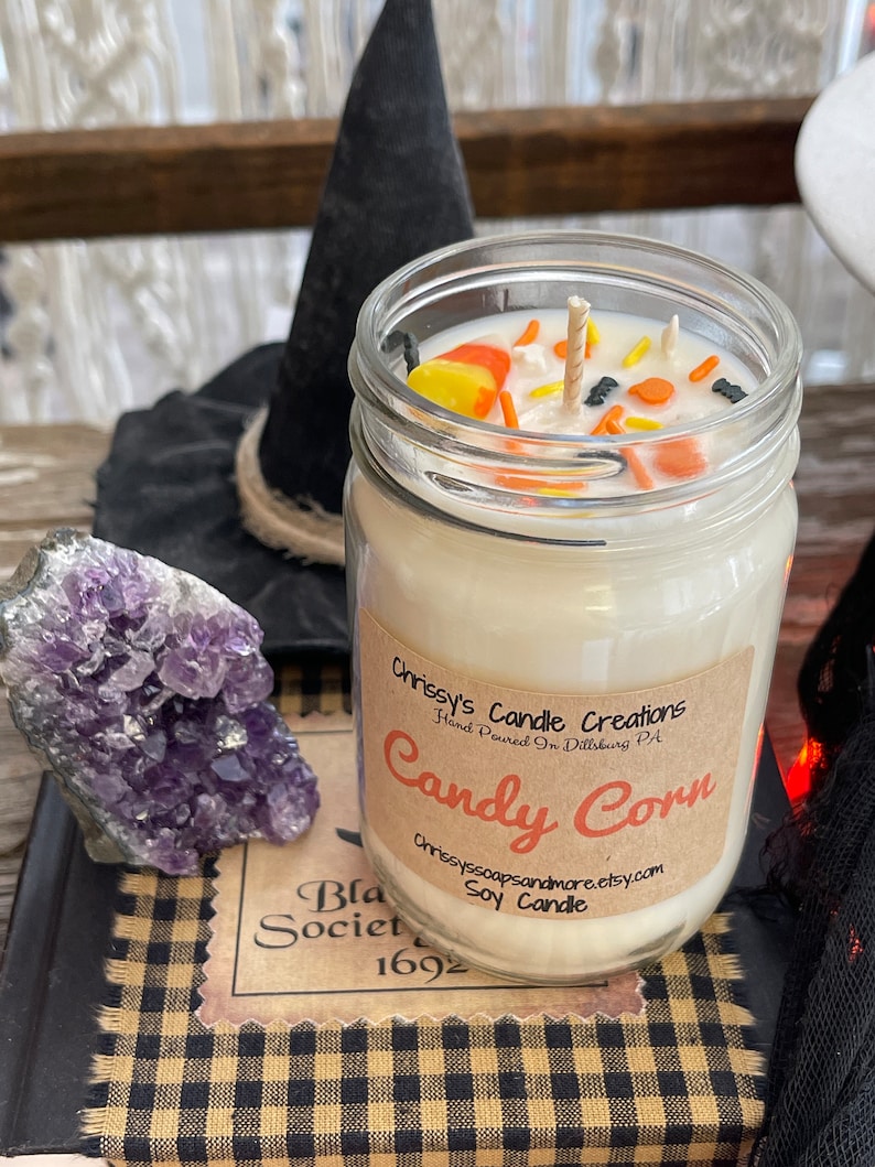 May include: A glass jar candle with a white soy wax base and a candy corn scent. The jar is topped with a wooden wick and decorated with candy corn pieces and sprinkles. The label reads "Chrissy's Candle Creations Hand Poured in Dillsburg PA Candy Corn Chrissyssoapsandmore.etsy.com Soy Candle".