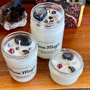 May include: Three glass jars filled with white wax and black stones. The jars are labeled "Moon Magic Soy Candle Infused with Healing Crystals and Herbs".