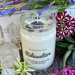 May include: A white soy candle in a clear glass jar with a label that reads "Relaxation Soy Candle infused with Healing Crystals and Herbs". The candle is surrounded by lavender flowers, clear quartz crystals, and green plants.
