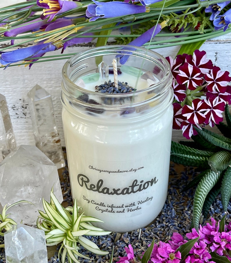May include: A white soy candle in a clear glass jar with a label that reads "Relaxation Soy Candle infused with Healing Crystals and Herbs". The candle is surrounded by lavender flowers, clear quartz crystals, and green plants.