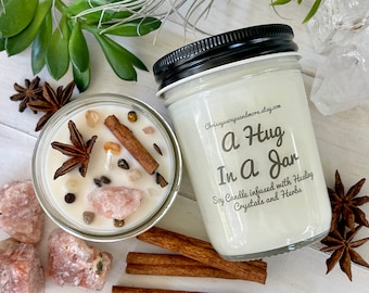 May include: Two glass jar candles with black lids. One candle has the text "A Hug In A Jar" and is decorated with crystals and herbs. The other candle is filled with wax, star anise, cinnamon sticks, and crystals.