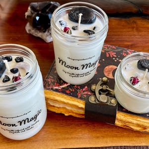 May include: Three white soy candles in glass jars with black stones and dried flowers inside. The candles are labeled "Moon Magic Soy Candles infused with Herbs, Crystals and Herbs".