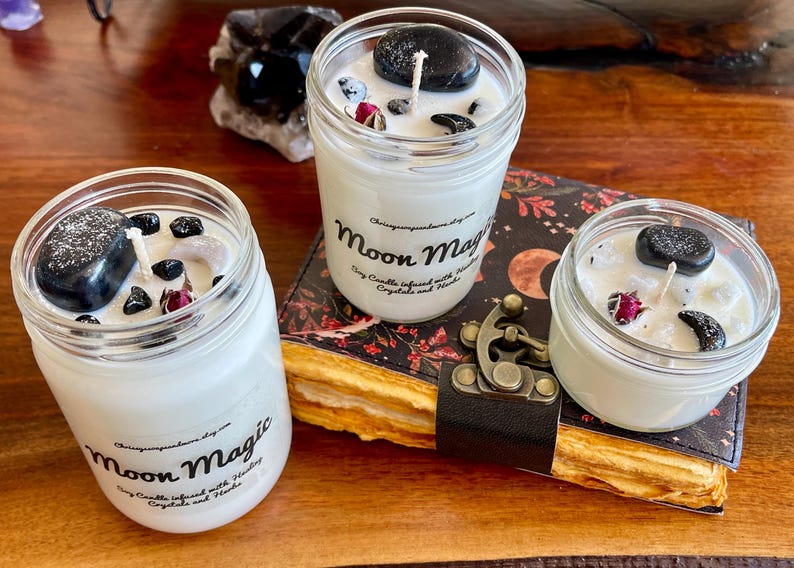 May include: Three white soy candles in glass jars with black stones and dried flowers inside. The candles are labeled "Moon Magic Soy Candles infused with Herbs, Crystals and Herbs".