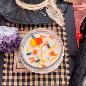 May include: A white candle with a candy corn, black bats, and orange pumpkins on top. The candle is in a clear glass jar with a black and tan checkered background.