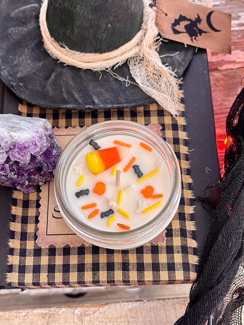 May include: A white candle with a candy corn, black bats, and orange pumpkins on top. The candle is in a clear glass jar with a black and tan checkered background.