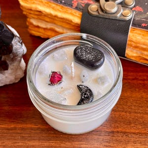 May include: A white candle in a glass jar with a black moon, black stone, and a dried red rose. The candle is surrounded by small white stones.