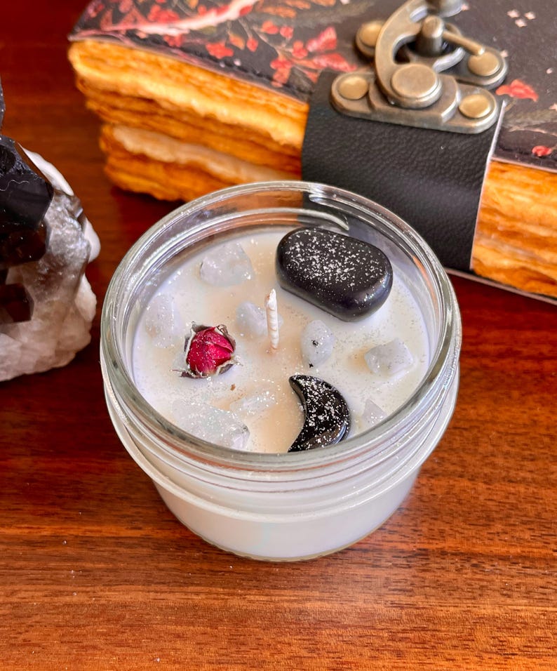 May include: A white candle in a glass jar with a black moon, black stone, and a dried red rose. The candle is surrounded by small white stones.