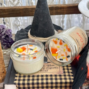 May include: Two Halloween-themed candles in glass jars with lids. One candle is topped with white wax and decorated with orange and yellow candy corn, black sprinkles, and a white wick. The other candle has a white wax top with a label that reads "Candy Corn". Both candles are sitting on a book with a black and white checkered pattern.