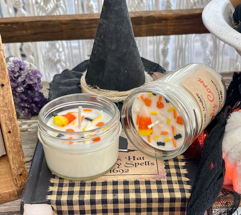 May include: Two Halloween-themed candles in glass jars with lids. One candle is topped with white wax and decorated with orange and yellow candy corn, black sprinkles, and a white wick. The other candle has a white wax top with a label that reads "Candy Corn". Both candles are sitting on a book with a black and white checkered pattern.
