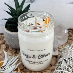 May include: A white soy candle in a glass jar with a sage and citrus scent. The candle is infused with healing crystals and herbs. The jar has a label that says "Sage & Citrus" and "Soy Candle infused with Healing Crystals and Herbs".