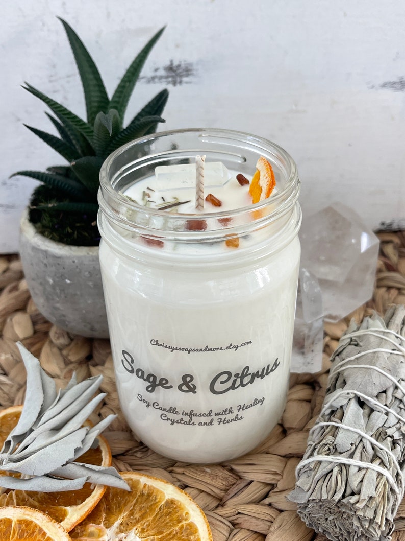 May include: A white soy candle in a glass jar with a sage and citrus scent. The candle is infused with healing crystals and herbs. The jar has a label that says "Sage & Citrus" and "Soy Candle infused with Healing Crystals and Herbs".